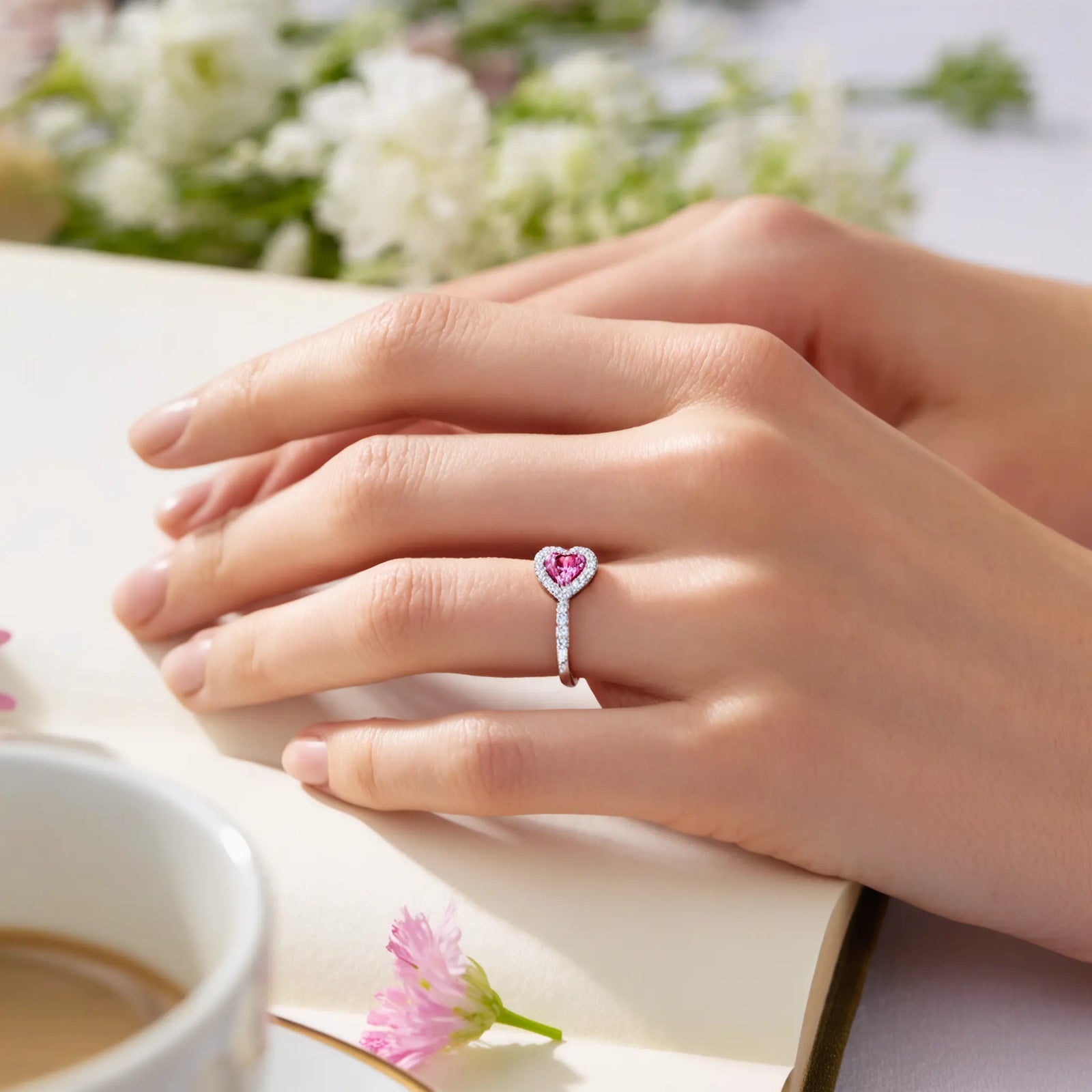 Woman's hand wearing a silver ring with a pink heart-shaped gemstone, resting on a book with flowers and coffee